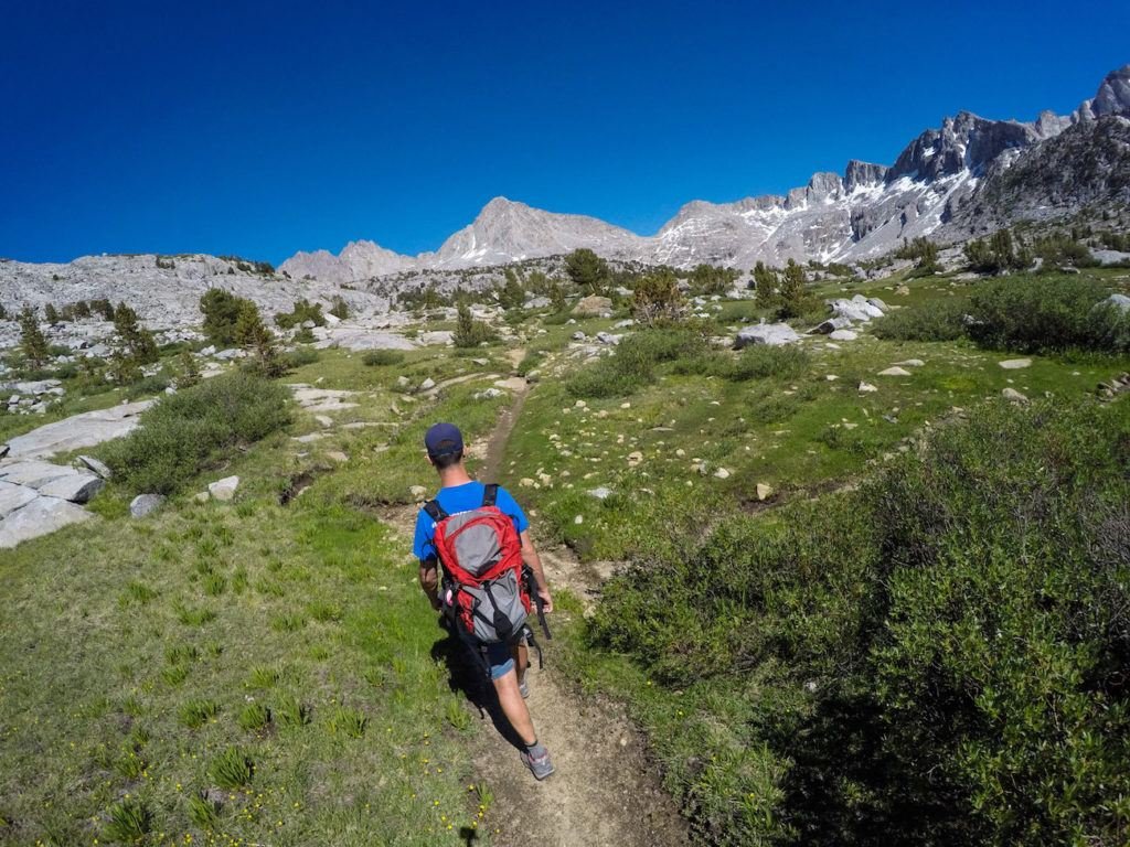 Man on hiking trail surrounded by green meadows and tall rocky mountains in backcountry of Eastern Sierras in California